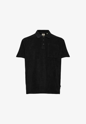 Mand iført sort kortærmet polo shirt, sorte tætsiddende bukser med hænderne i lommerne, og hvide sneakers, stående mod en ensfarvet baggrund.