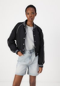 Chaqueta bomber negra con puños y cuello de rayas blancas, usada sobre un sudadera gris. Pantalones cortos de mezclilla azul claro. Textura de tela suave.
