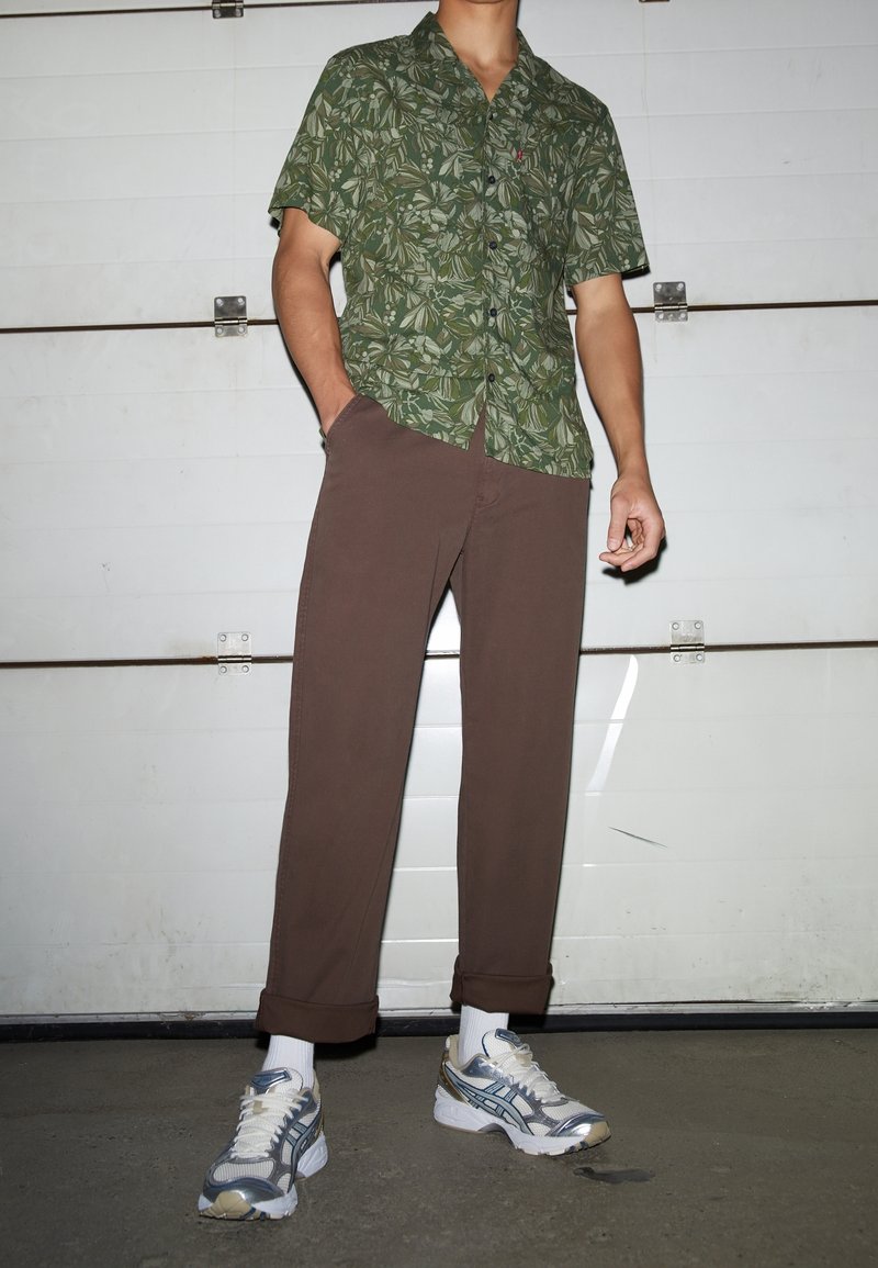 Grön mönstrad kortärmad skjorta med mörka löv, i kombination med bruna byxor och grå sneakers. Uppvikta ärmmuddar på byxorna.