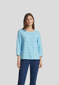Helles blaues Oberteil mit einem strukturierten wellenförmigen Muster, mit langen Ärmeln und einem runden Ausschnitt. Kombiniert mit dunkellblauen Jeans.