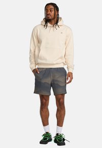 Beige Kapuzenpullover mit Fronttasche, kombiniert mit grauen Shorts mit einem Bergmuster. Dazu werden schwarze Sneaker und weiße Socken getragen.