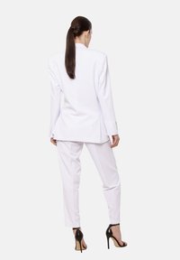 Kobieta z długimi ciemnymi włosami w białym garniturze i czarnych sandałach na wysokim obcasie stoi tyłem na jednolitym tle.