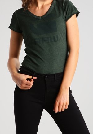 Kobieta ubrana w ciemnozielony dopasowany T-shirt z dekoltem w serek z wyblakłym logo oraz czarne spodnie z wysokim stanem, trzymająca ręką za pas.