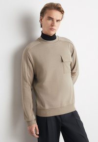 Felpa beige con colletto a girocollo, ampi polsini a costine e una piccola tasca sul petto. Realizzata in morbido tessuto, abbinata a pantaloni neri.