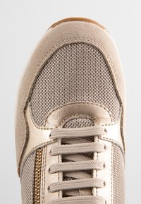 Scarpa sportiva beige realizzata in suede e materiali a rete, con chiusura con lacci, dettagli metallici e una superficie testurizzata.