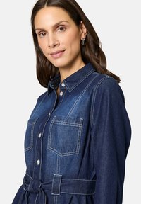 Denim overhemdjurk in donkerblauw met twee voorzakken, knoopsluiting en een ceintuur in de taille. Kenmerkt zich door contrasterende witte stiksels en een kraag.