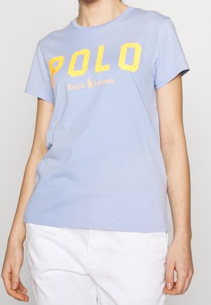 Persoon die een lichtblauw T-shirt met korte mouwen draagt met "POLO Ralph Lauren" in grote gele letters en een witte broek.