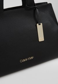 Sac à main Calvin Klein en cuir noir texturé avec logo doré et étiquette rectangulaire en or suspendue à la poignée.