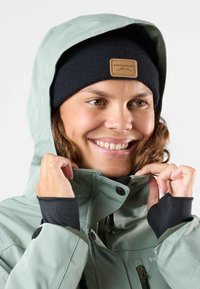 Light green waterproof jacket with a hood, zippered front, and black wrist cuffs. Black knit beanie with a tan leather logo patch.