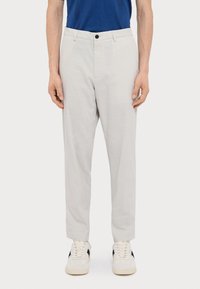 Pantaloni grigi chiari, design aderente, realizzati in cotone, con chiusura frontale a bottone, tasche laterali e indossati con sneakers bianche.