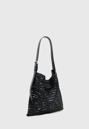 Μαύρη τσάντα tote πλεκτή με ρυθμιζόμενο λουράκι ώμου από δέρμα και ασημένια αγκράφα, εμφανίζεται σε απλό ανοιχτό γκρι φόντο.