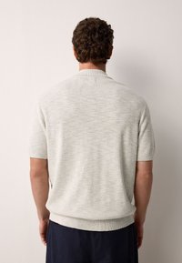 Hellgrauer Strickpullover mit kurzen Ärmeln und strukturiertem Muster, mit einem gerippten Ausschnitt und Saum, getragen über einer dunklen Hose.