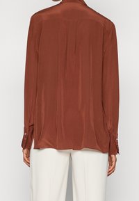 Blouse en satin marron à manches longues avec une coupe décontractée, poignets à boutons et un subtil pli dans le dos, associée à un pantalon crème.