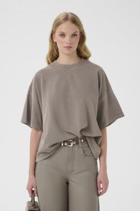 Ξανθιά γυναίκα που φοράει υπερμεγέθη φανέλα σε χρώμα ταουπ, μπασμένη σε αντίστοιχο παντελόνι ψηλής μέσης με ζώνη, κρατώντας μια υφασμάτινη τσάντα στο πλάι της.