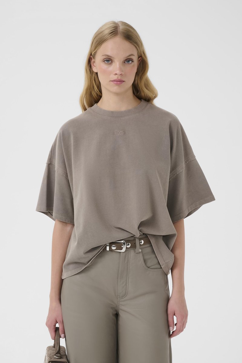 Femme blonde portant un t-shirt taupe surdimensionné rentré dans un pantalon taille haute assorti avec une ceinture, tenant un sac à main texturé à son côté.