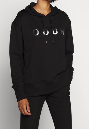 Persoon draagt een zwarte hoodie met glanzende zilveren letters "HUGO", voorzak en trekkoorden, gecombineerd met een zwarte broek.