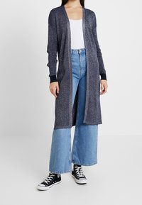Lång marinblå cardigan med metalltråd, öppen design, ribbade ärmar, ihop med högmidjade ljusblå jeans och svarta kanvas sneakers.