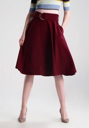 Femme portant une jupe trapèze bordeaux avec une large ceinture, associée à un haut rayé coloré et des escarpins beige, debout avec une main dans la poche.