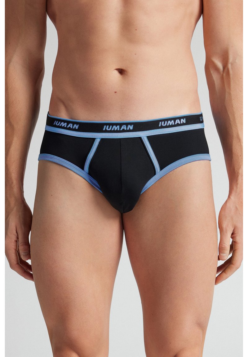 Mandlig overkrop iført sorte underbukser med blå kant og linning mærket "IUMAN", stående med armene afslappet ned langs siderne mod hvid baggrund.