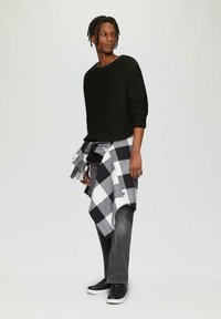 Schwarzer Strickpullover, graue Jeans und schwarze Sneakers. Ein schwarz-weiß kariertes Hemd ist um die Taille gebunden. Einfaches Design und Texturen.