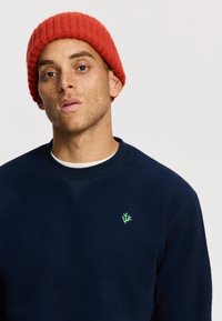Mornarsko moder sweatshirt z zelenim logotipom na levem prsnem predelu; v kombinaciji z svetlo rdečo rebrasto pleteno kapo. Gladka tekstura, sproščen oblikovanje.