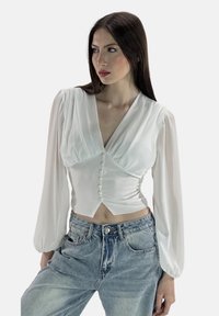 Blusa bianca con maniche lunghe a sbuffo e scollatura a V, caratterizzata da una chiusura con bottoni e un tessuto morbido e fluido. Abbinate a jeans a vita alta.
