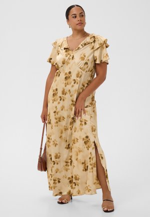 Kvinde iført en beige blomster-maxikjole med flæseærmer og sidelår med slids, der holder en brun håndtaske og har sorte åbne tå-hæle på.