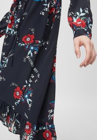 Blommig klänning i mörk marinblått tyg med röda och blå blommor, långa ärmar, rynkad fåll och en dragsko-detalj i midjan.