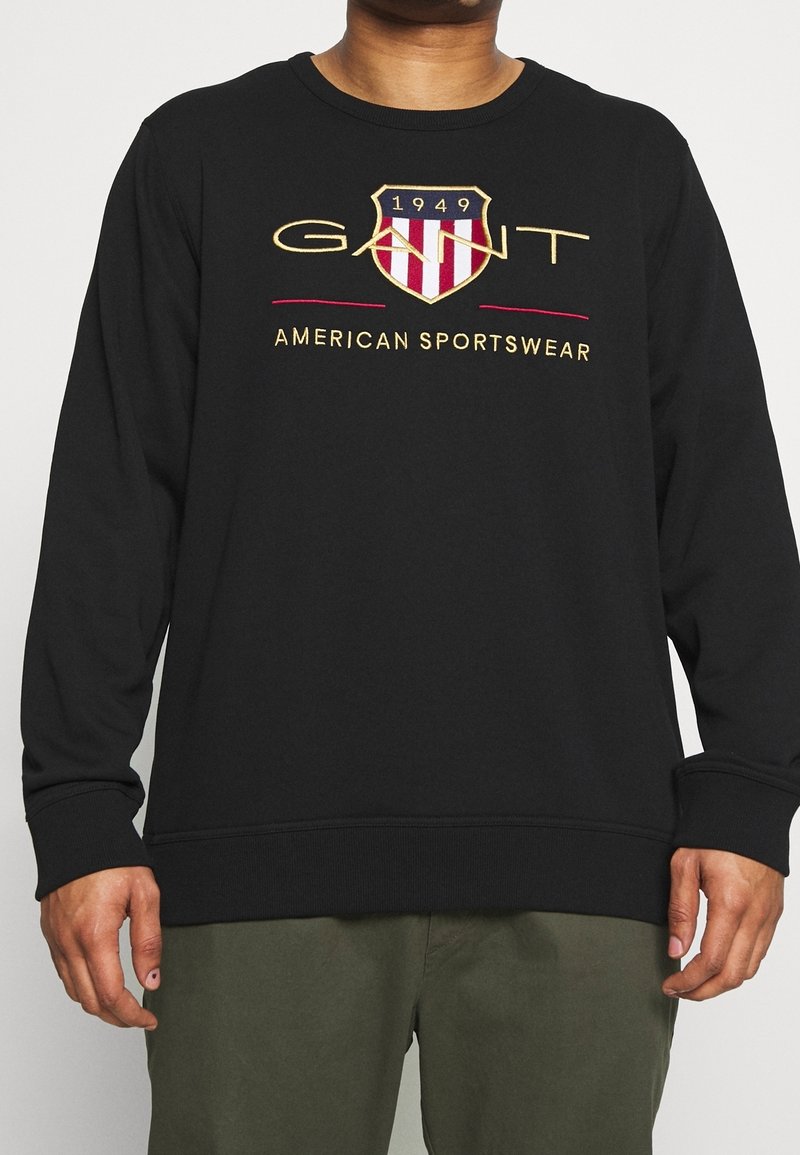 Personne portant un sweat-shirt noir avec le texte American Sportswear et le logo bouclier 1949, associé à un pantalon vert.