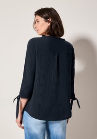 Blusa in blu navy, con texture, dal taglio dritto, maniche a tre quarti e dettagli con fiocco nei polsini. Il retro presenta una discreta piega.