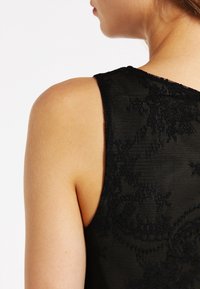 Plano detalle del hombro y cuello de una mujer que lleva una blusa negra de encaje sin mangas, mostrando la textura de la piel y los detalles de la tela.