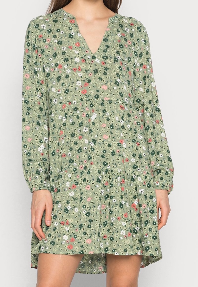 Femme portant une robe verte à fleurs à manches longues avec un col en V et une taille froncée, debout devant un fond clair uni.
