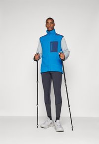Blue fleece vest with a gray long-sleeve base layer, black pocket, paired with gray leggings and white hiking shoes, holding hiking poles.