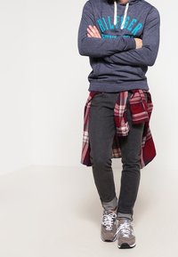 Sudadera marina con un logo turquesa, jeans gris oscuro y zapatillas grises. Camisa de cuadros rojos atada a la cintura como un detalle de acento.