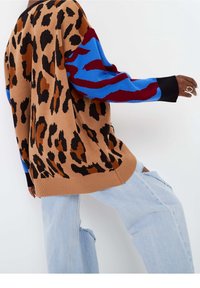 Luipaardprint trui in bruin en zwart met blauwe en bordeauxrode mouwen, gecombineerd met lichtblauwe versleten jeans. Gebreide stof, casual ontwerp.