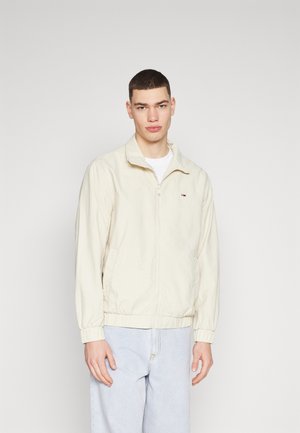 Chaqueta ligera de color beige claro con cuello alzado, cremallera frontal y puños elásticos; presenta un pequeño logo en el lado izquierdo del pecho.