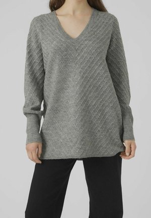Kvinde iført langærmet grå strikket V-hals sweater med diagonalt ribbet mønster, kombineret med sorte bukser, stående mod en ensfarvet baggrund.