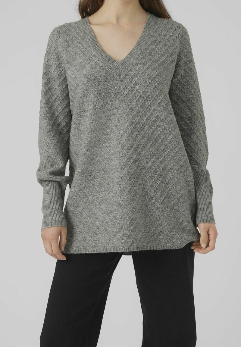 Femme portant un pull gris à manches longues en maille à col en V avec un motif côtelé diagonal, associé à un pantalon noir, debout devant un fond uni.