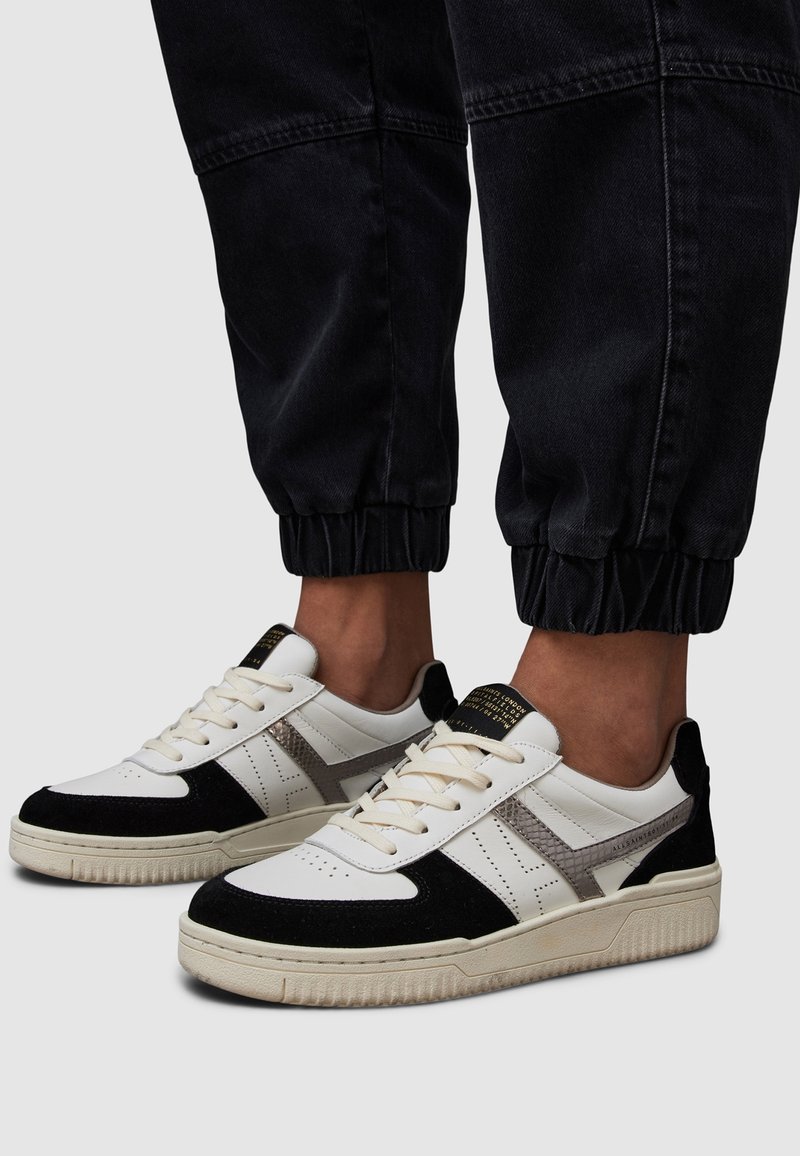 Baskets basses blanches, noires et grises sur une personne portant un pantalon noir retroussé sur un fond uni.