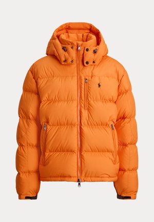Personne portant une doudoune orange, une casquette verte, des lunettes de soleil foncées, un pull violet, un jean bleu et des chaussures marron, debout les mains dans les poches.