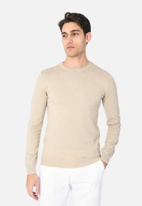 Maglione beige a maniche lunghe in tessuto morbido con scollatura rotonda e polsini a costine, abbinato a pantaloni bianchi. Design semplice e aderente.
