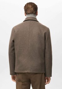 Brauner Mantel mit Struktur und hohem Kragen, mit einer Rückennaht und leicht strukturierten Schultern, getragen über hellen Hosen.