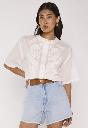 Donna che indossa una camicetta bianca ricamata con maniche corte, pantaloncini chiari in denim e una cintura marrone con fibbia rotonda.