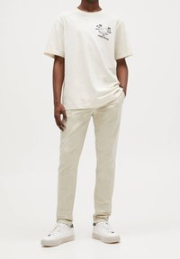 Persona che indossa pantaloni beige chiari, sneakers bianche e una maglietta avorio con un piccolo logo "The Chill Frog" sul petto.