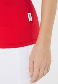 Rotes, figurbetontes Top aus glattem Stoff, mit einem seitlichen Etikett mit dem Aufdruck "SENSES". Weiße Hose mit einem klassischen Schnitt ist daneben sichtbar.