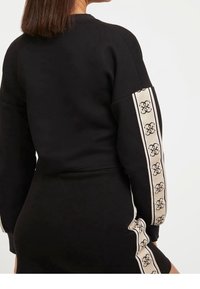 Kobieta w czarnym, długim rękawie topie i spódnicy w kremowe paski, z czarnymi splecionymi inicjałami na rękawach i boku spódnicy.