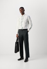 Λευκό πουκάμισο με πράσινο αφηρημένο σχέδιο, μαύρη ζώνη, σκούρες παντελόνες και παπούτσια, κρατώντας μια μαύρη δερμάτινη τσάντα, στέκεται σε λευκό φόντο.