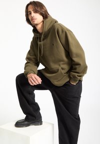 Olivengrøn hættetr�øje med snøre, frontlomme og logo. Parret med sorte jeans og mørke sko, stående på en hvid pedestal.