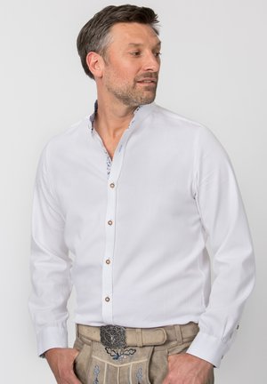 Man wearing white buttoned shirt with mandarin collar and beige traditional embroidered lederhosen standing with one hand in pocket, looking sideways.