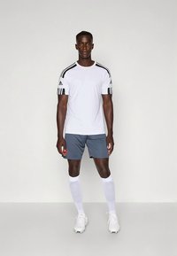 Witte atletische T-shirt met zwarte strepen op de schouders, gecombineerd met grijze korte broek. Model draagt witte sokken en voetbalschoenen en staat neutraal.